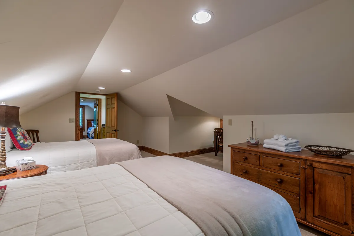 A comfortable bedroom with two beds covered in crisp white bedding, a wooden dresser, a table lamp with a decorative shade, and an open door leading to an adjoining room. The room feels inviting and warm, suitable for families or friends.