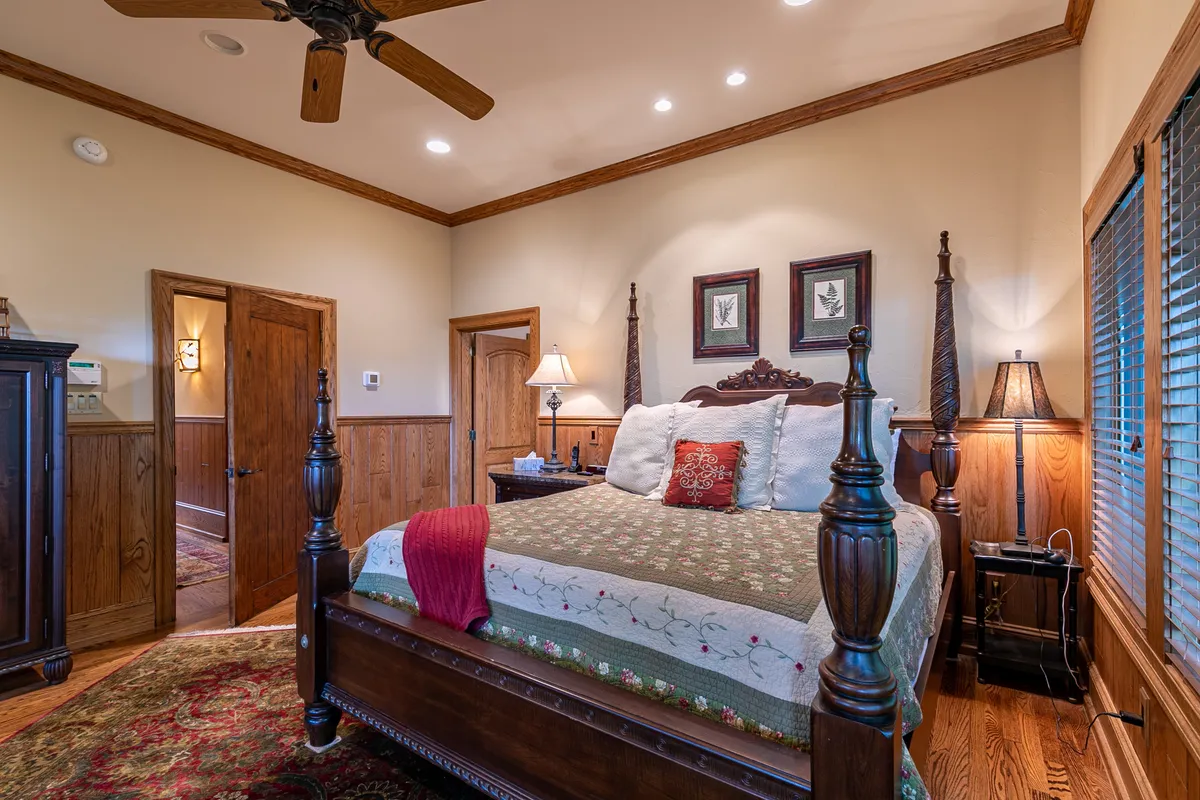 A serene bedroom with an ornate bed, decorative pillows, substantial wooden furniture, and artwork on the walls. The space is softly lit, exuding comfort and calm, ideal for relaxation or reading.
