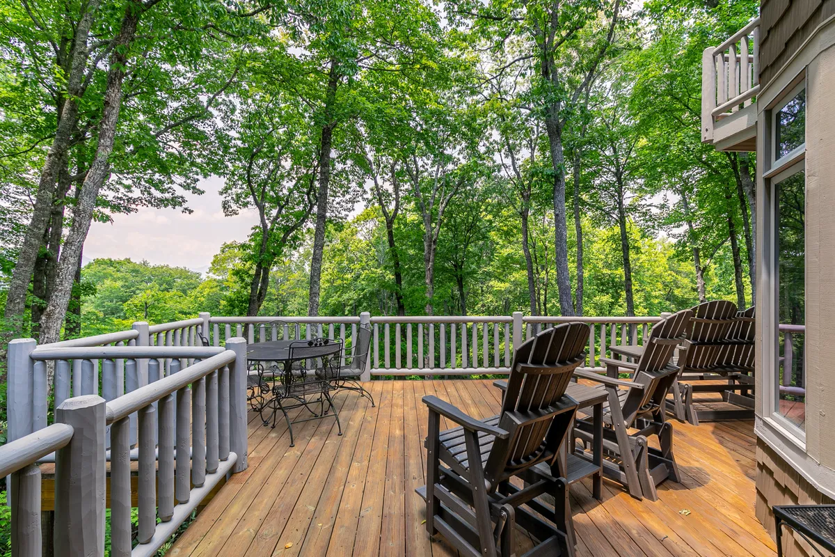 A spacious wooden deck bordered by Adirondack chairs, an outdoor dining table, and lush surrounding trees. The area offers serene views of nature, perfect for group gatherings and morning coffee rituals. The layout invites socializing while immersed in mountain tranquility.