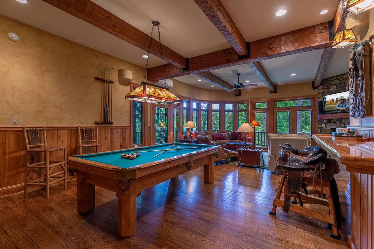 A spacious entertainment room featuring a Proline pool table set beneath exposed wooden beams, a cozy seating area by the fireplace, and a distinctive decorative saddle that hints at unique saddle-bar stools. Large windows offer natural light, and the space is designed for social gatherings and relaxation.
