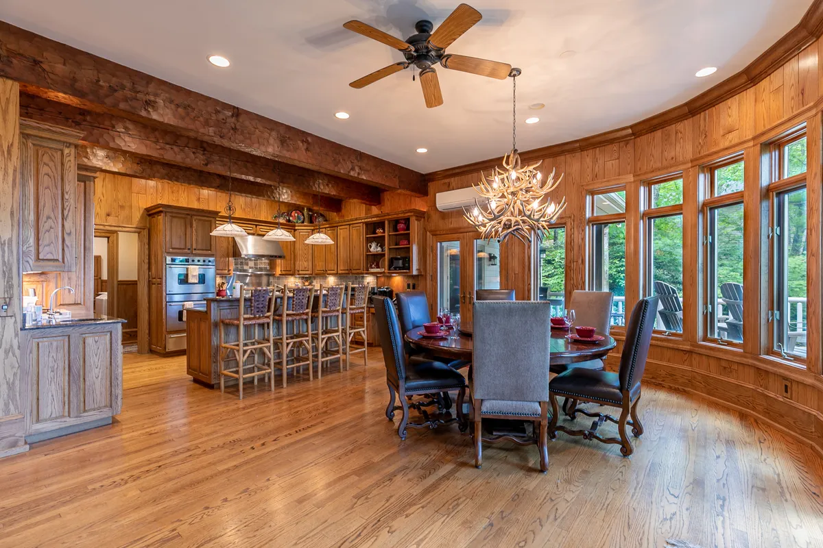An elegant dining area featuring a large wooden table for eight, with a beautiful chandelier overhead and an adjoining kitchen island with bar stools. The space is bathed in natural light from the expansive curved windows, offering views and a welcoming ambiance.
