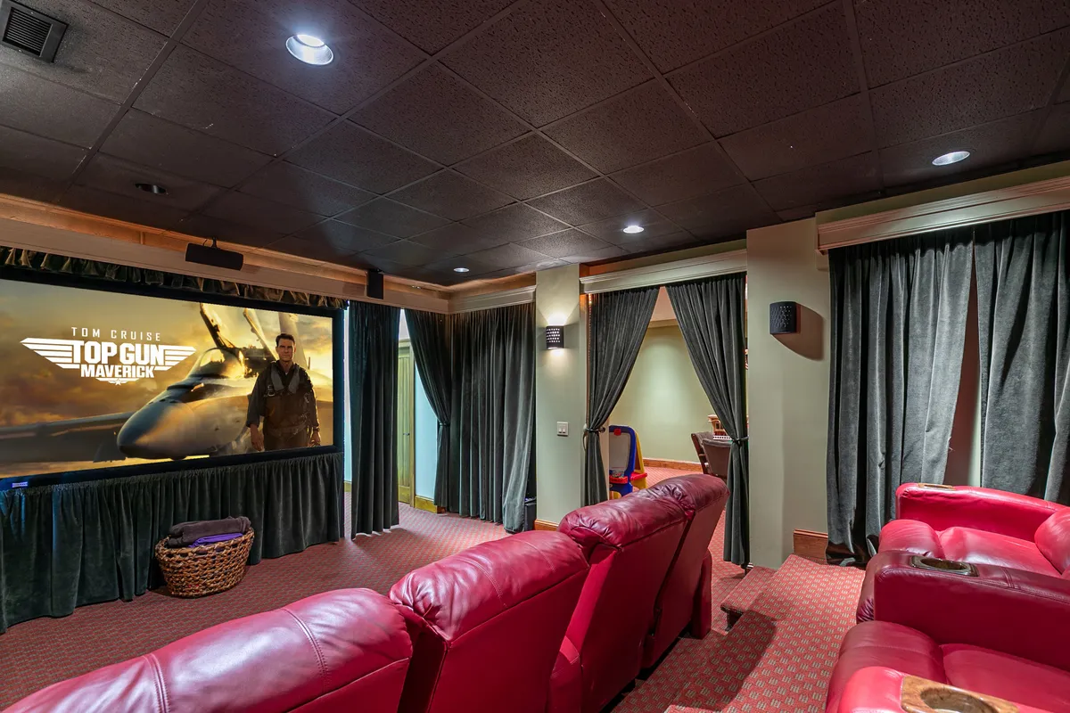 A private, in-home theater featuring a large movie screen displaying 'Top Gun: Maverick,' flanked by red leather seating and plush curtains. The space exudes entertainment and comfort, ideal for all-weather enjoyment with family and friends.
