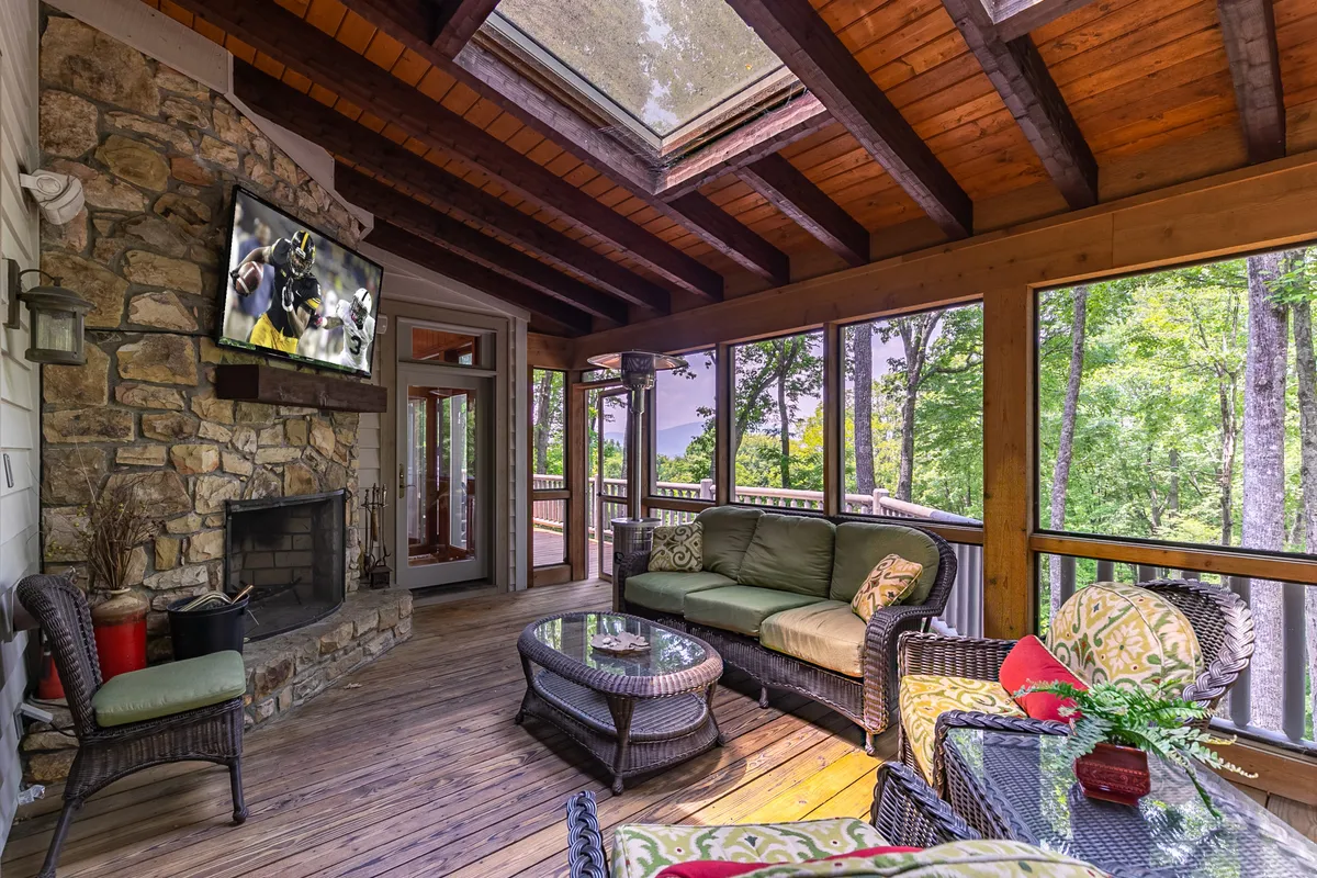 A three-season screened deck with a stone fireplace, television, wicker furniture, skylight, and large windows that open the room to the outdoors. The setup is ideal for relaxing, watching a game, or enjoying the ambiance of an indoor-outdoor space.