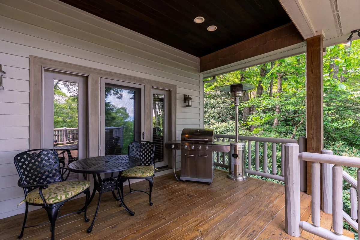 An inviting exterior patio featuring a round dining table with black chairs and green cushions, a grill, a patio heater, and a wooden railing. The setup creates an ideal environment for outdoor dining, evening barbecues, and relaxed social gatherings against a backdrop of nature.