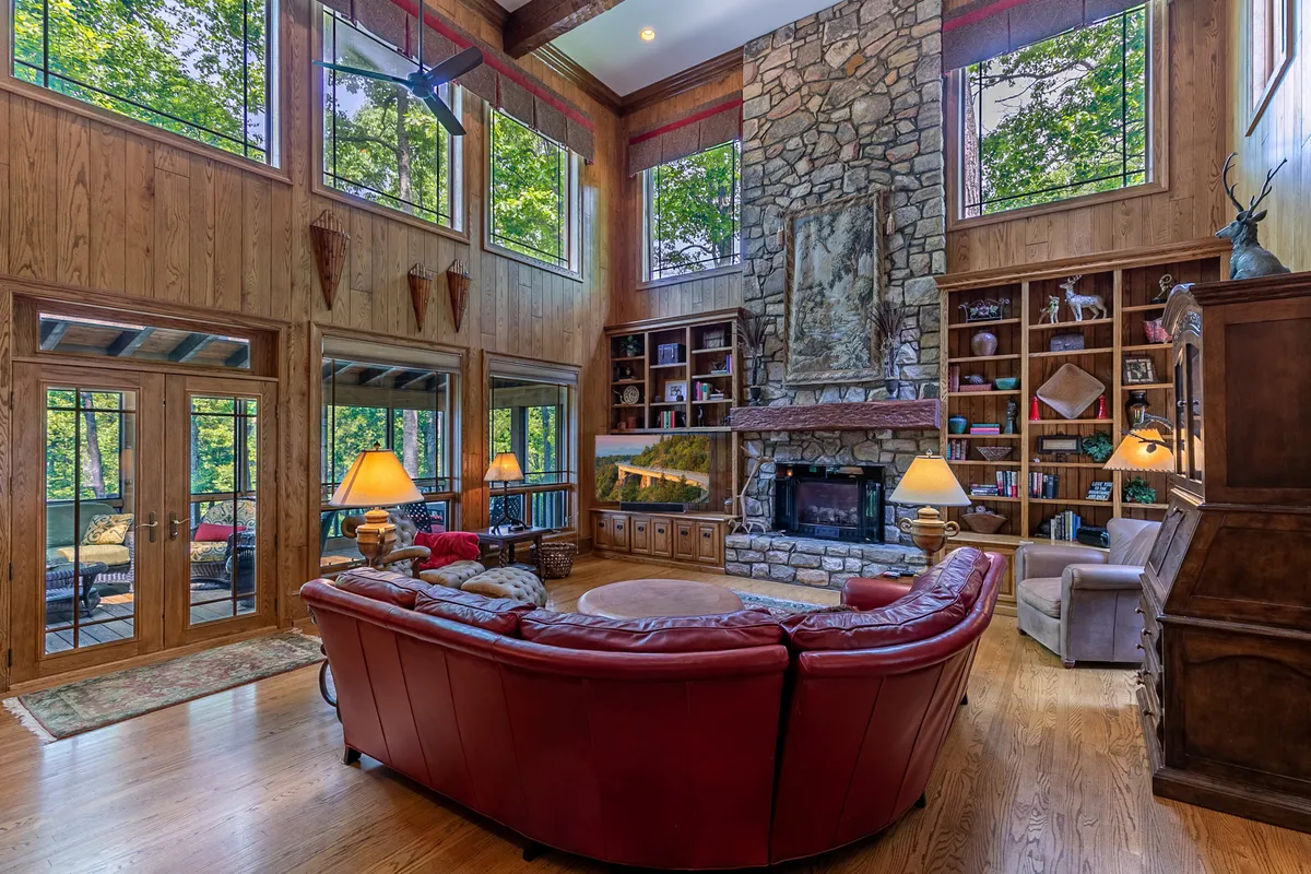 This image showcases an inviting living room with a dramatic stone fireplace, expansive windows letting in natural light, red leather seating, and wooden bookshelves lining the walls. The atmosphere is warm and welcoming, ideal for family gatherings or quiet relaxation by the fire.