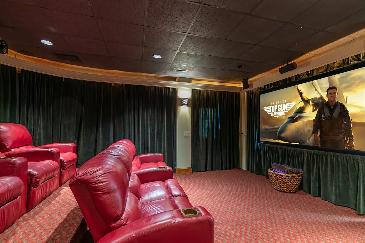 A luxurious cinema-style theater room featuring plush red leather recliners, a massive movie screen displaying 'Top Gun: Maverick,' and dark velvet curtains for an authentic cinematic experience. The setup invites guests to gather for private movie nights or themed screenings.