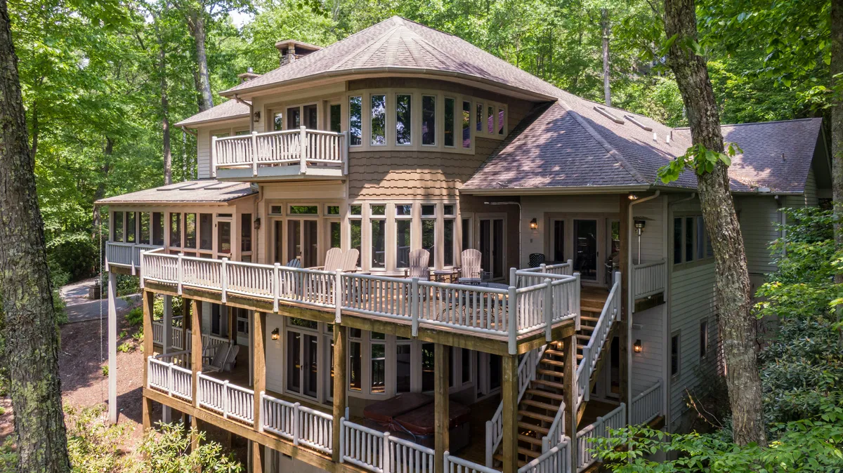 The hero image showcases the stunning exterior of a luxurious mountain home with a spacious wooden deck and multiple balconies. Large windows frame forested views, and inviting outdoor furniture hints at spaces perfect for dining or relaxing. The home is nestled among mature trees, capturing the tranquil ambiance of Boone, North Carolina, with the fading golden light of sunset enhancing its warmth and appeal.