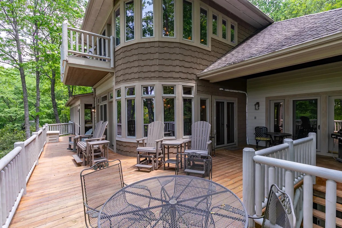 The image presents a serene deck area with seating, a round table, and wooden railings. Large windows from the house look onto the deck, which is enveloped by greenery, suggesting a direct connection to the outdoors for dining, relaxing, or wildlife watching.