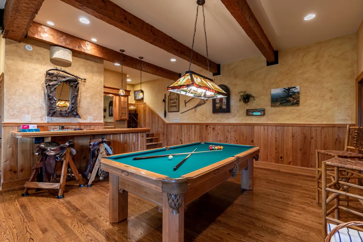 A cozy indoor recreation room with a pool table, a stained glass lamp overhead, and a wooden bar area. The space is designed for socializing, playing games, and enjoying drinks in a relaxed mountain setting.
