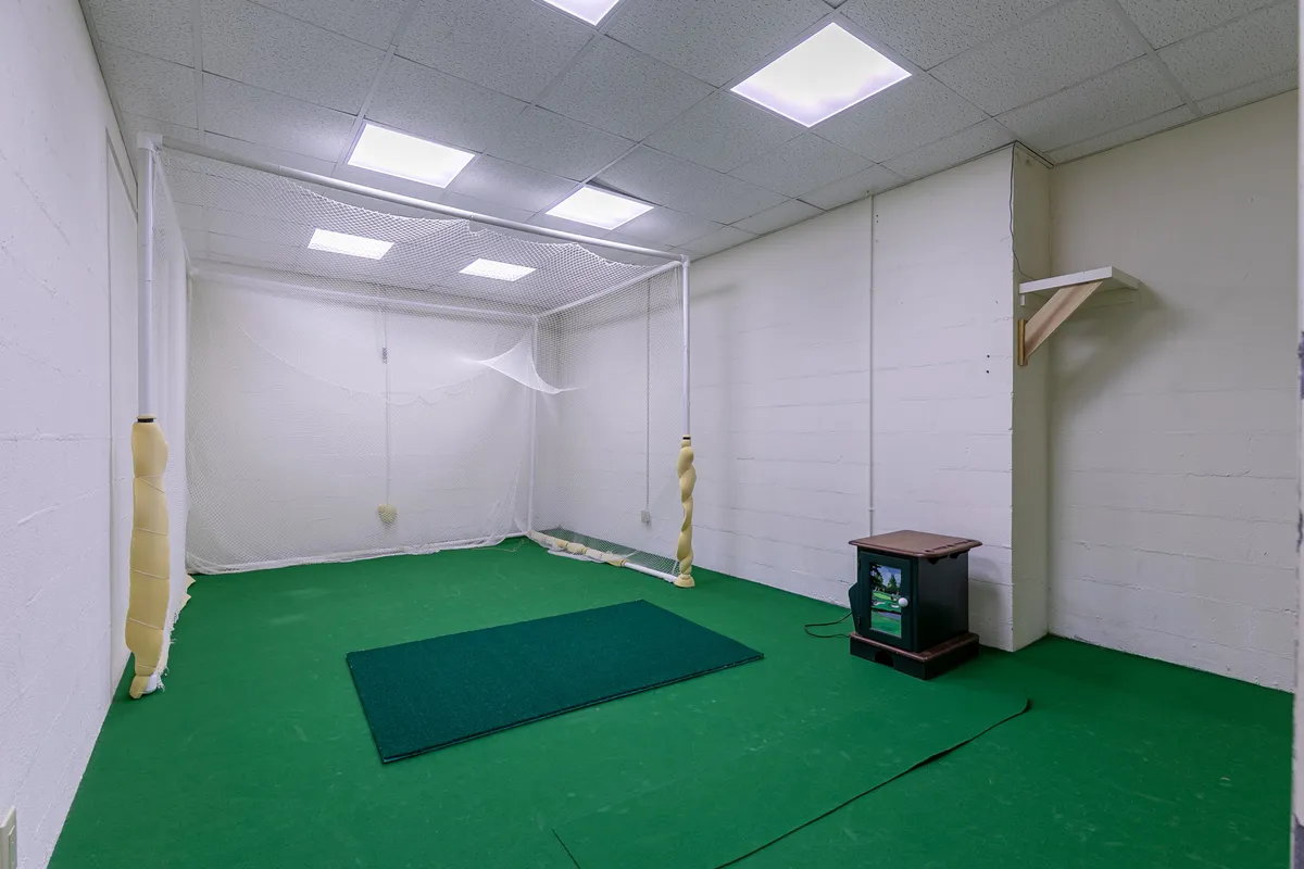 A golf practice room equipped with golf netting, a putting mat, and a small table. The focused layout provides an inviting space to hone golf skills, echoing a driving range experience within the home.