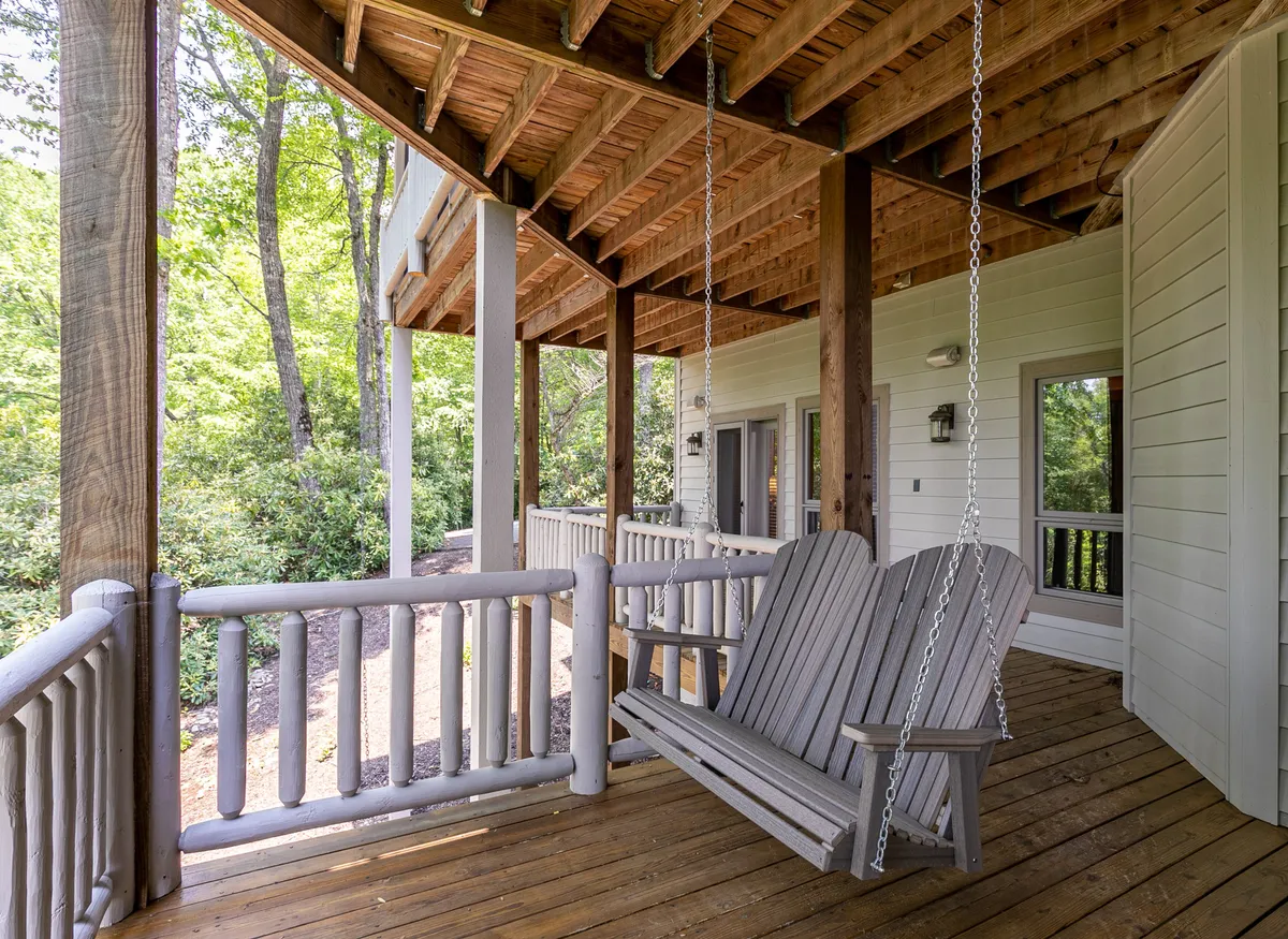 A serene exterior scene featuring a wooden swing seat on a deck, bordered by a sturdy wooden railing and surrounded by lush greenery. The tranquil setting invites guests to relax, read, or simply enjoy the sights and sounds of nature throughout the changing seasons.